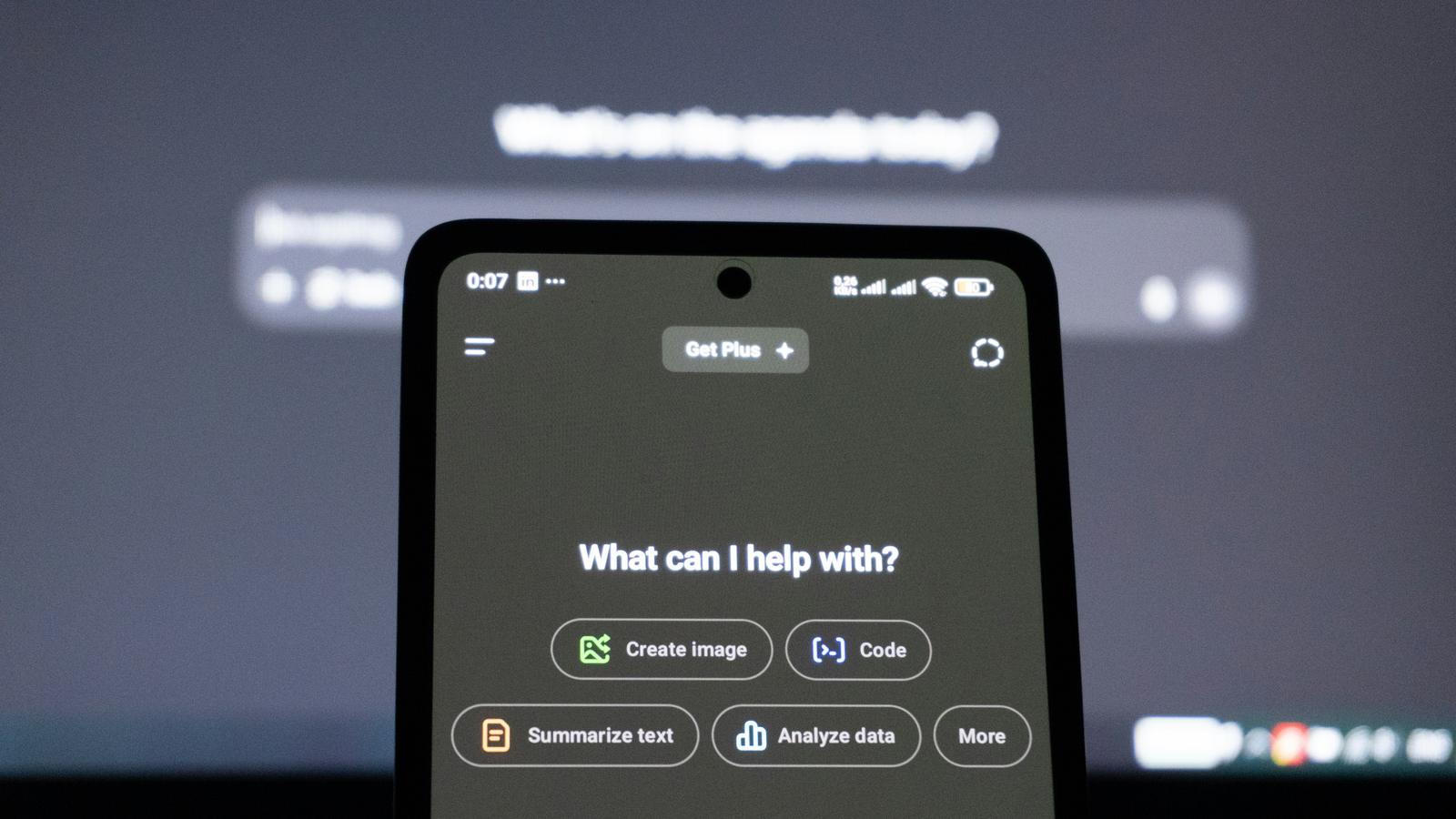 A smartphone showing an AI assistant's start screen, a computer screen blurred behind it