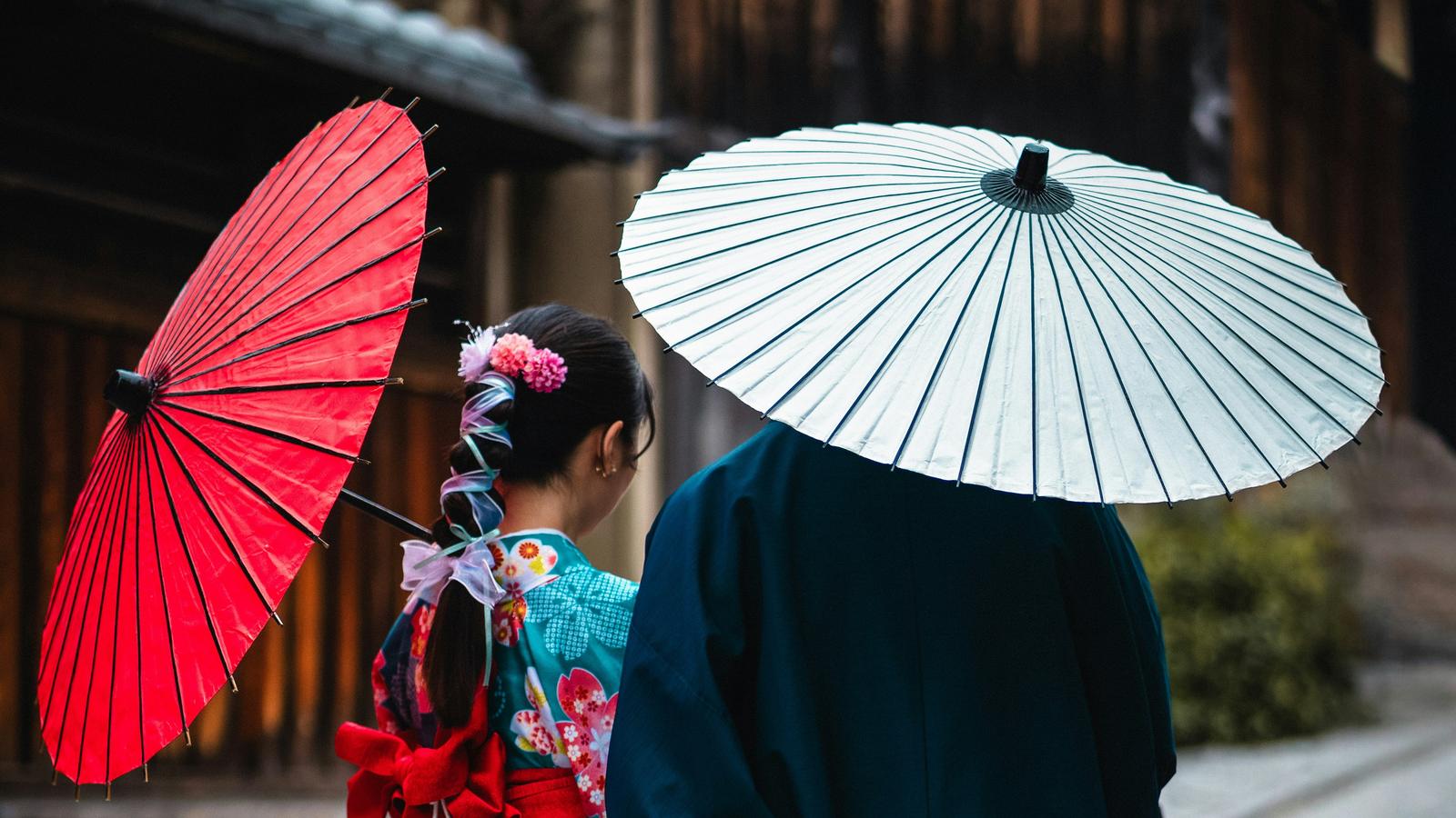 Two people in kimono standing side by side on a stone lane in Kyoto, holding red and white paper umbrellas
