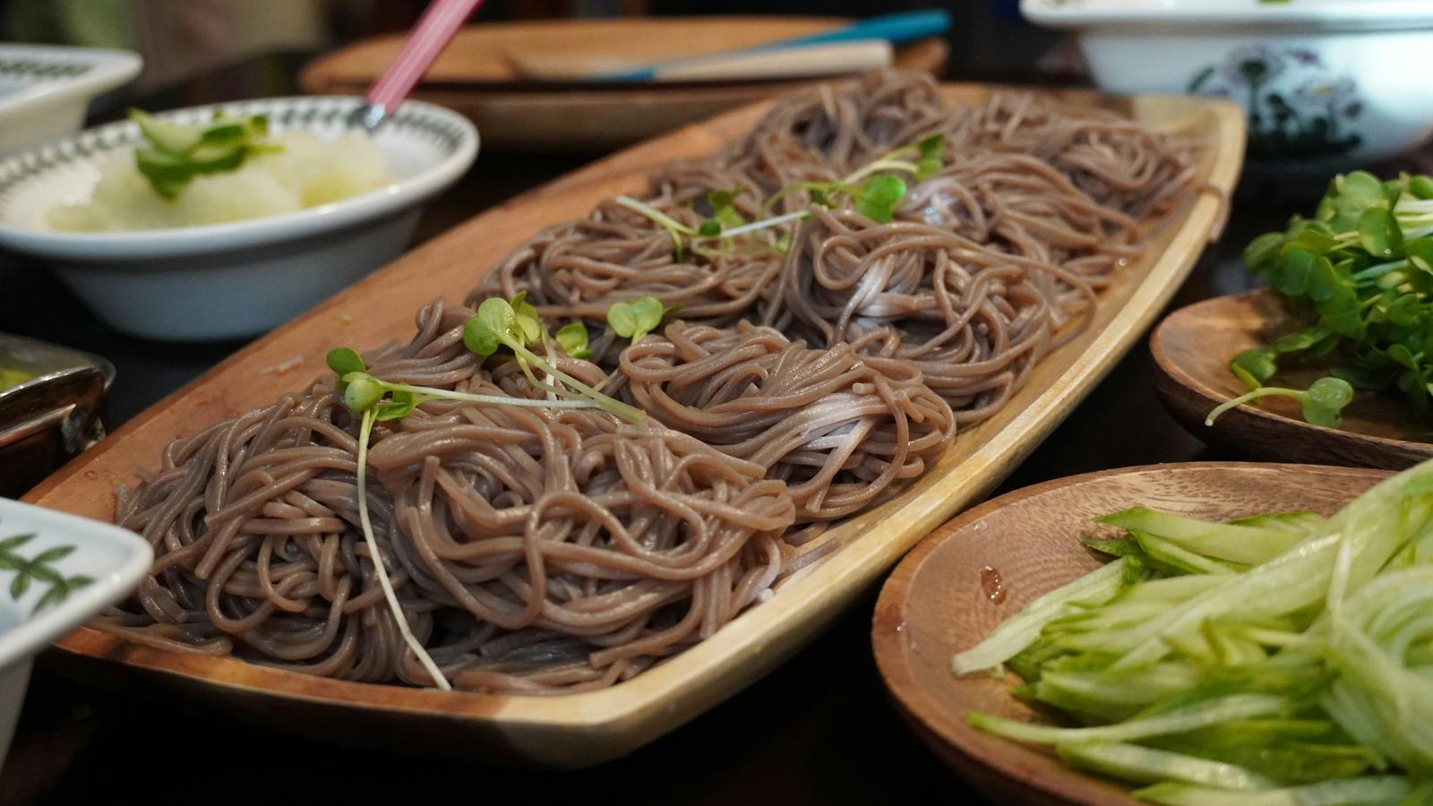 Cold buckwheat soba noodles served on a wooden tray, with a bowl of dipping sauce and shredded vegetables beside it