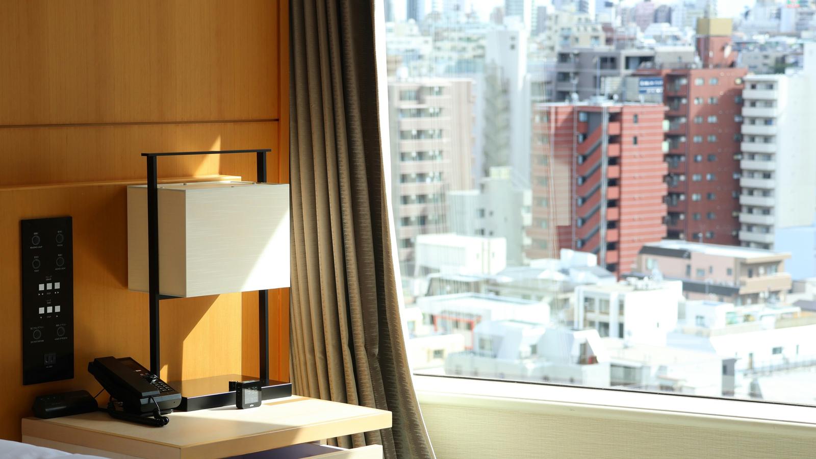 A bedside lamp and curtain beside a tall hotel window, with morning light on the apartment blocks of Tokyo below