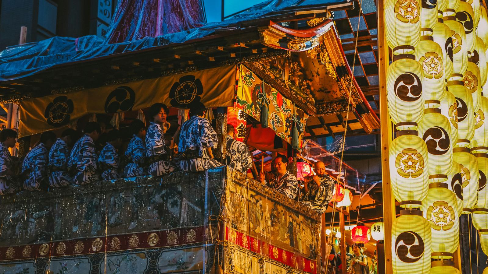 Musicians in yukata on a decorated festival float in Kyoto, beside a column of paper lanterns at dusk