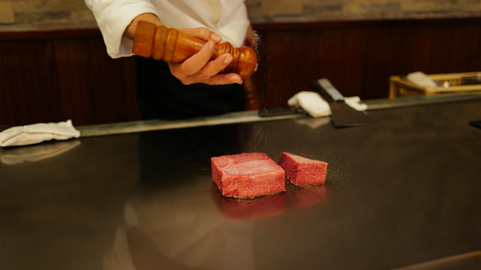 A chef at a teppanyaki griddle seasoning two thick cuts of beef