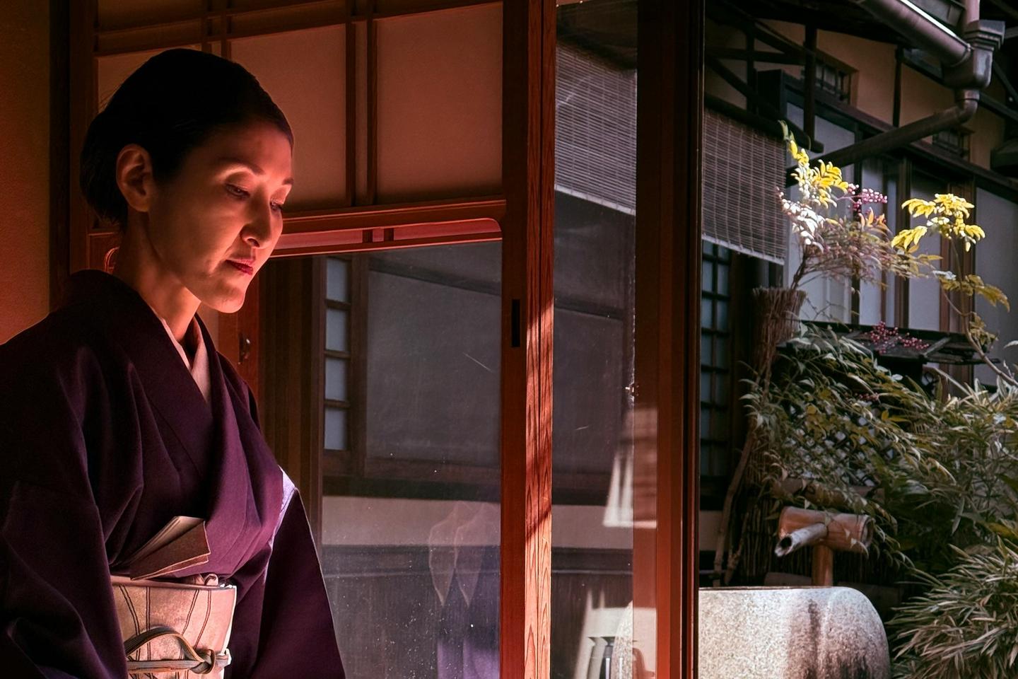 A woman in a dark kimono kneeling in a tea room by an open window, ladling hot water for tea