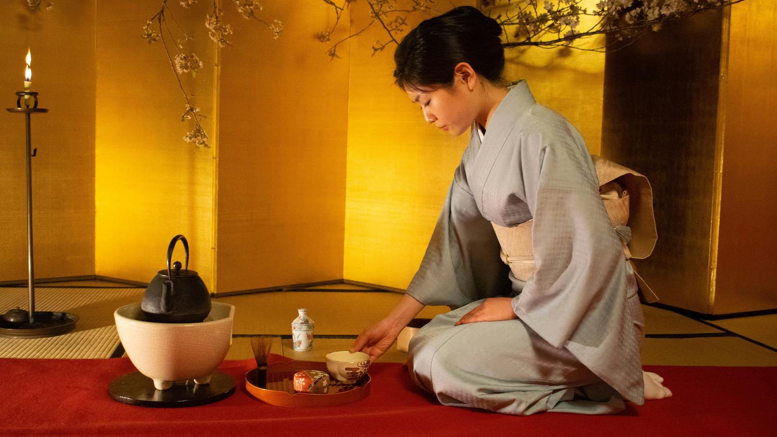 A woman in a pale kimono kneeling to prepare tea before a gold folding screen, with an iron kettle on a brazier beside her