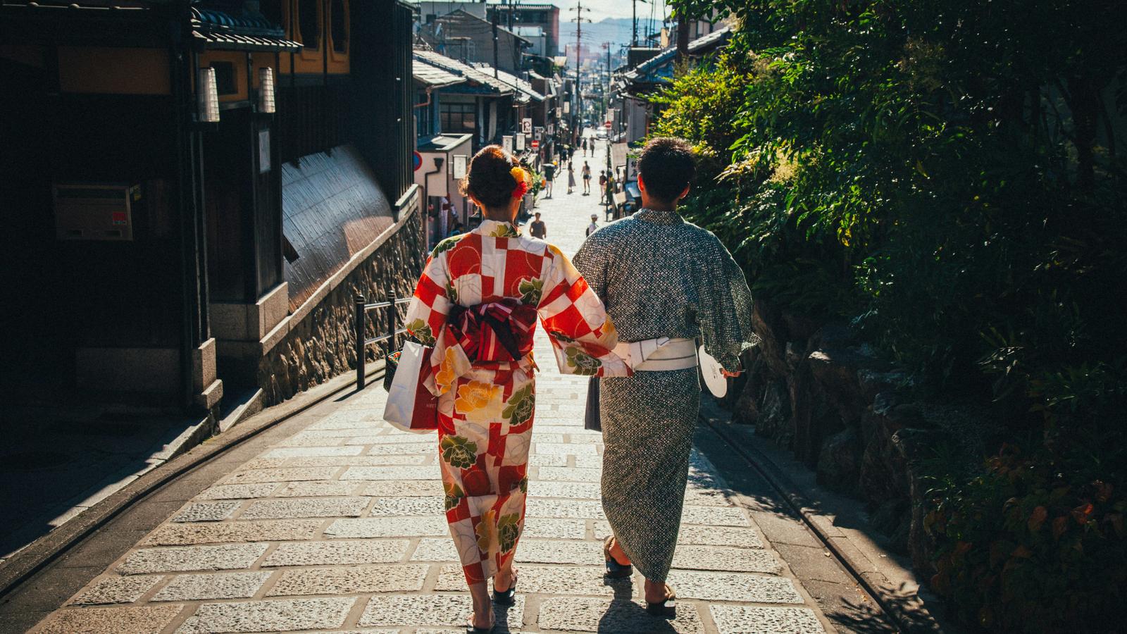 A couple in yukata walking side by side down a stone-paved street of old wooden houses in Kyoto, seen from behind