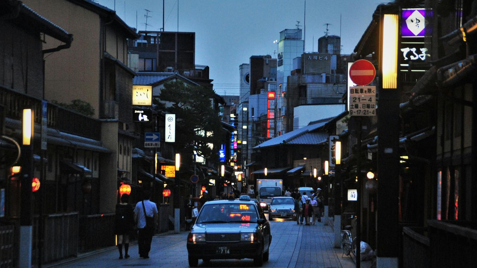 Early evening on a street of wooden townhouses in Gion, Kyoto, with lanterns lit and a taxi passing