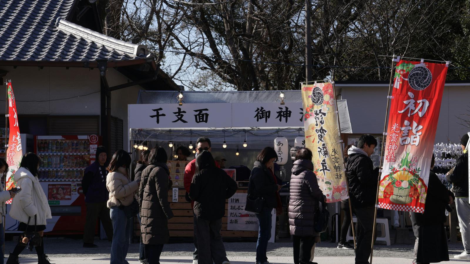 Visitors in winter coats queue at a shrine stall beneath red banners reading hatsumode, the first shrine visit of the New Year