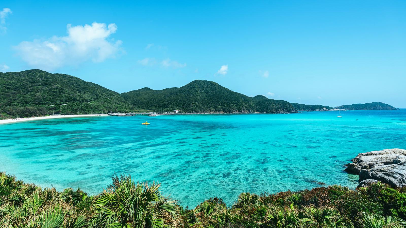 Clear turquoise water in a bay on Tokashiki Island, Okinawa, with a white beach below green hills