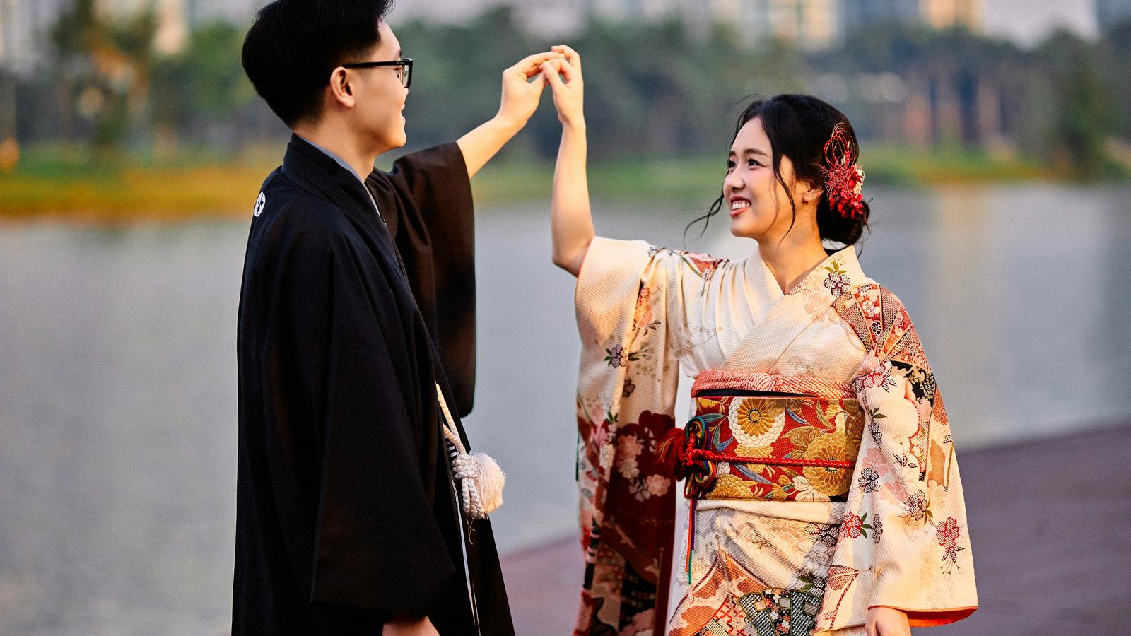 A couple in kimono holding hands beside a lake, smiling at each other