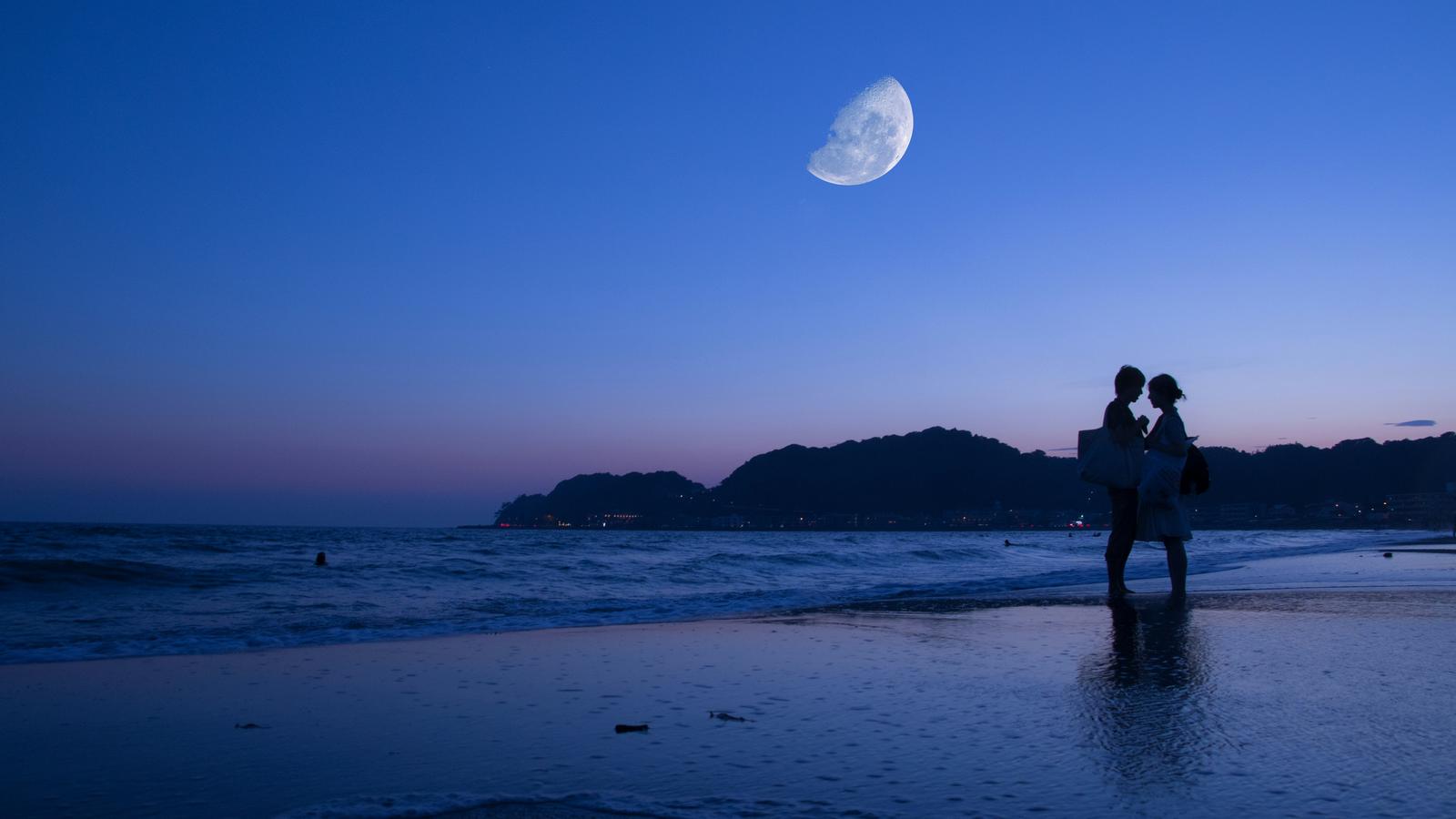 Two people embracing on a beach at dusk, with a half moon above the water