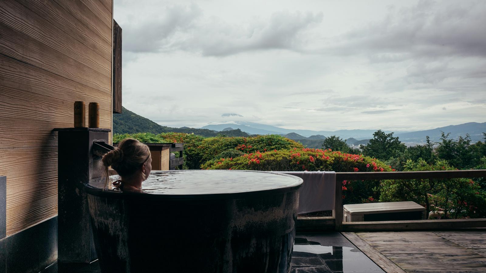 A guest seen from behind in a round private open-air hot spring bath on a terrace, looking out over green hills and distant mountains in Japan
