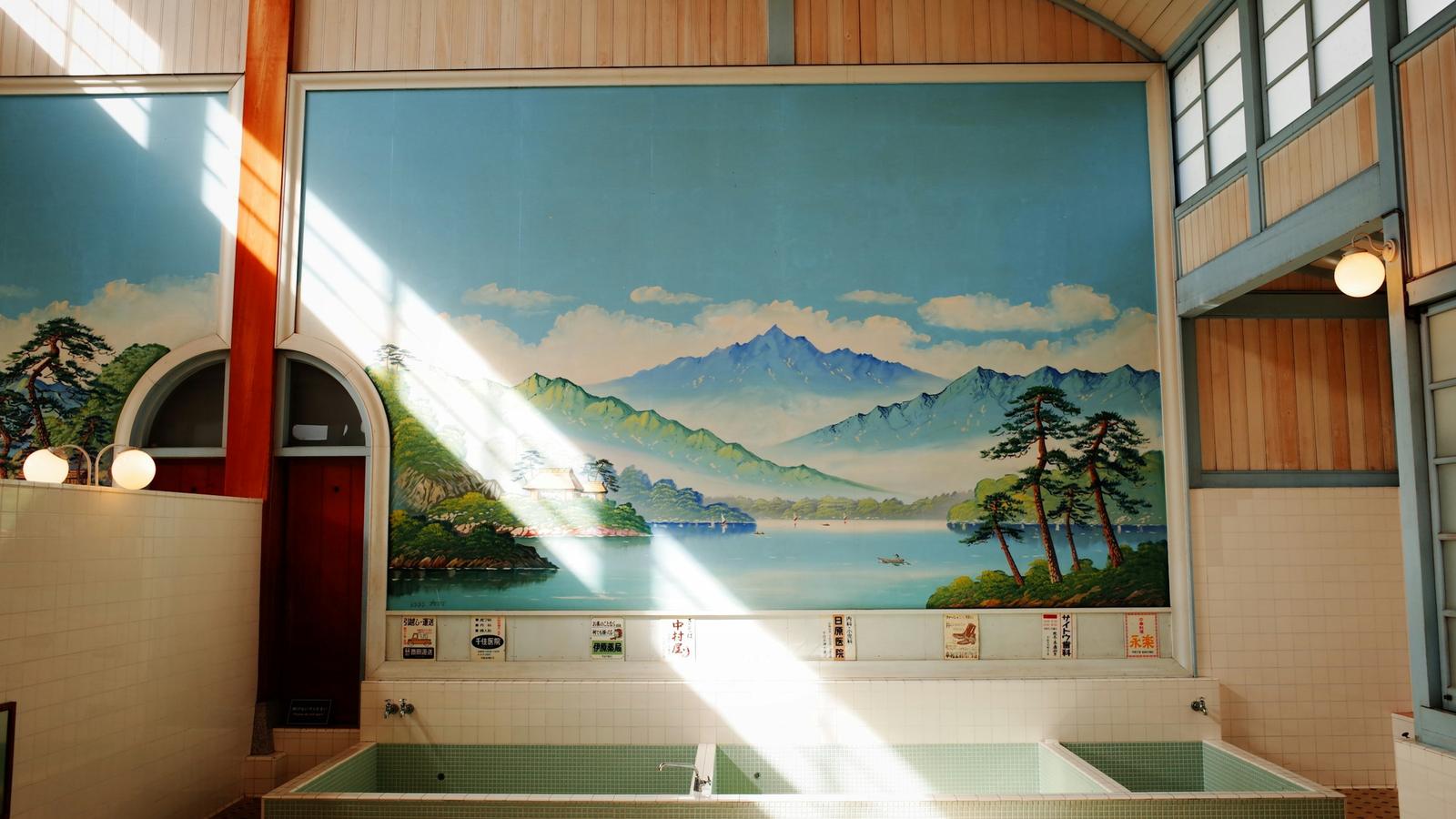 The bathing hall of a Japanese sento, with a painted mountain and lake mural above the baths and sunlight across the tiles