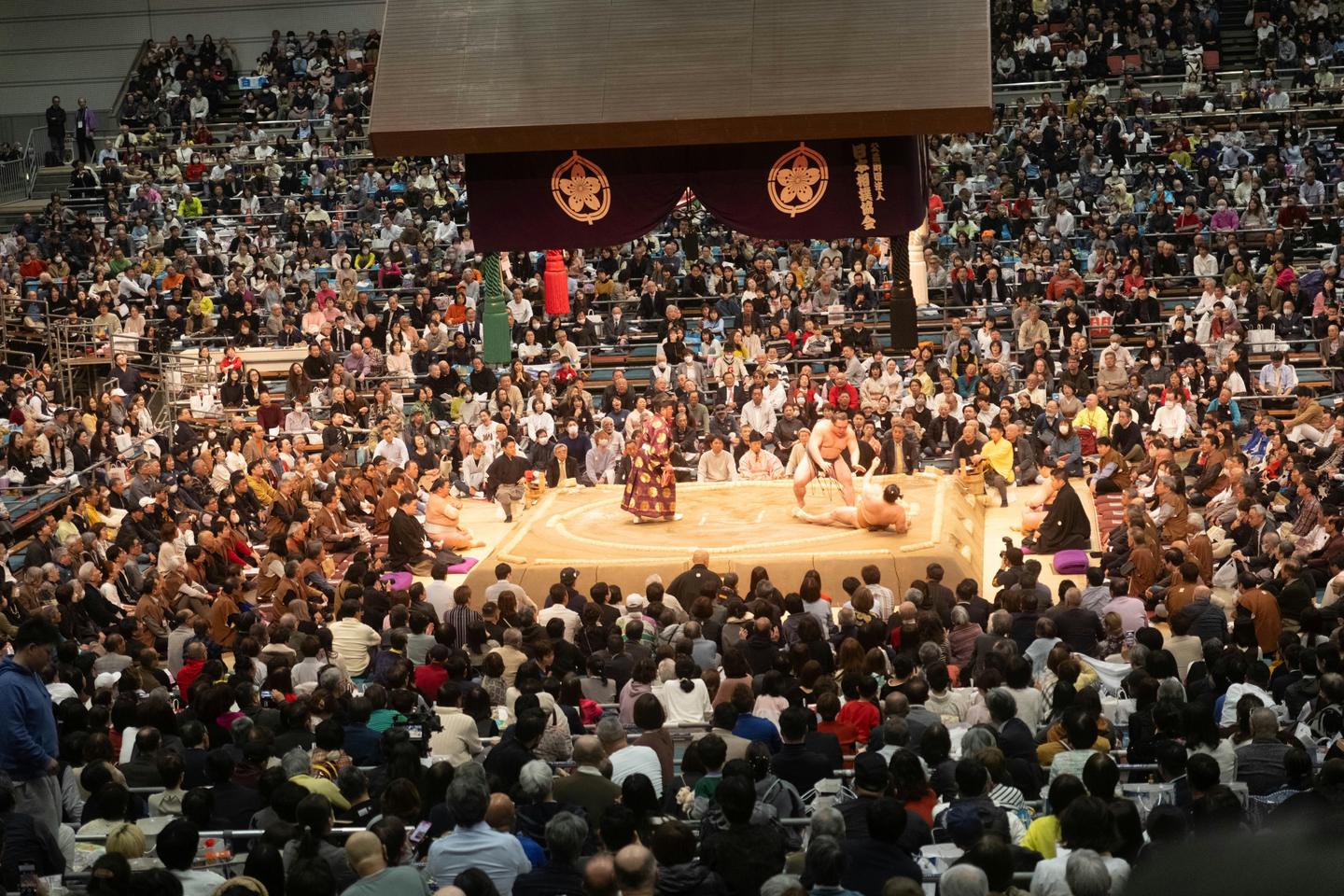 A bout ending at the edge of the ring as the referee looks on, with masu boxes and chair seats filled to the back of the arena