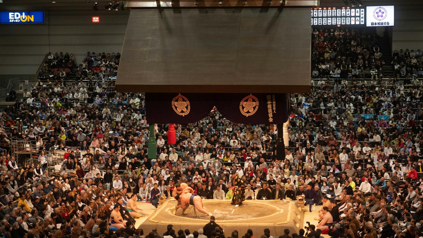 Two wrestlers grappling on the clay ring under the hanging roof at the March tournament in Osaka, with the hall full around them