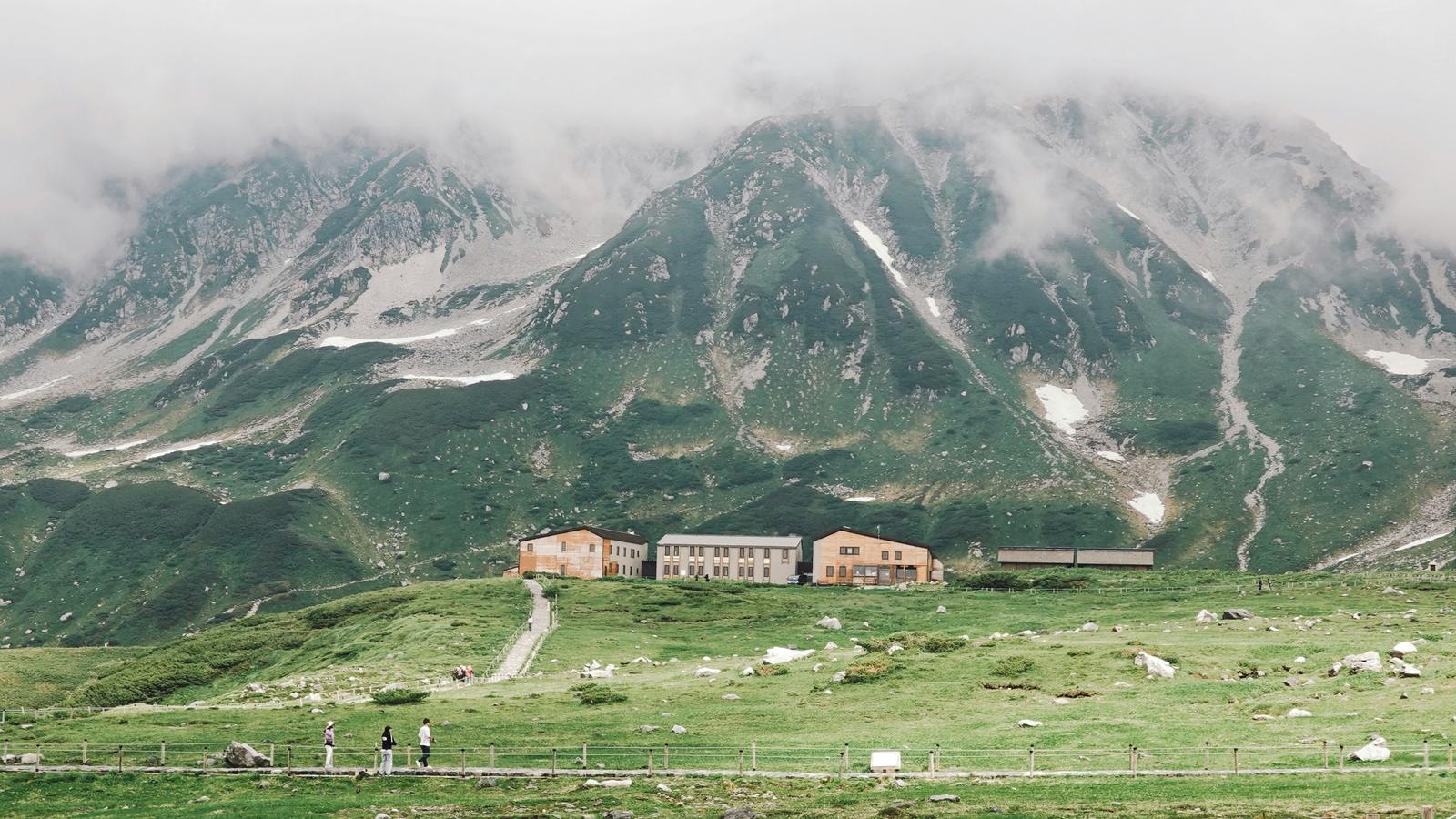 Mountain lodges stand on the green Murodo plateau below the Tateyama range, its upper slopes in cloud.