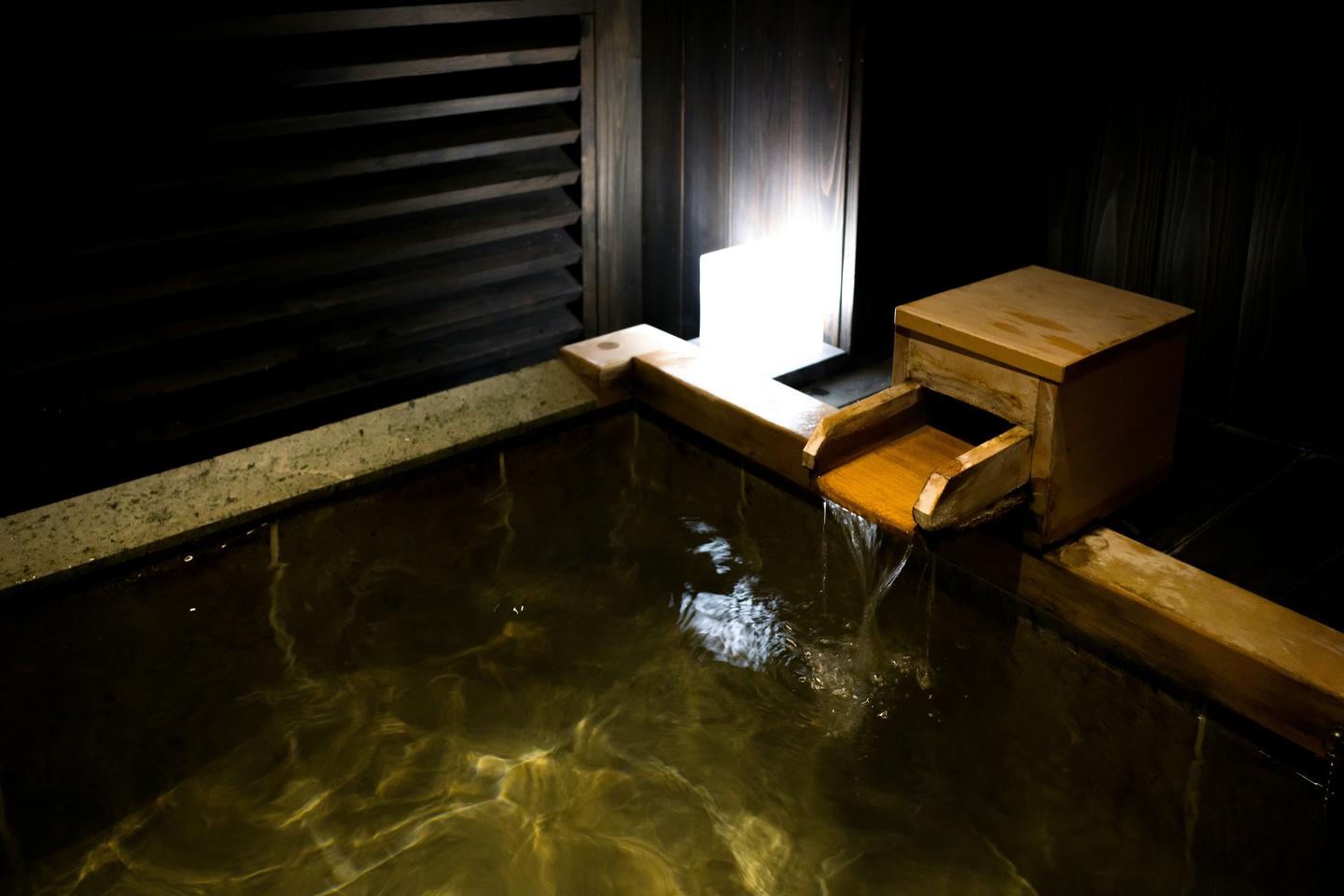 Hot spring water pouring from a wooden spout into a stone and timber bath lit low in a dark room