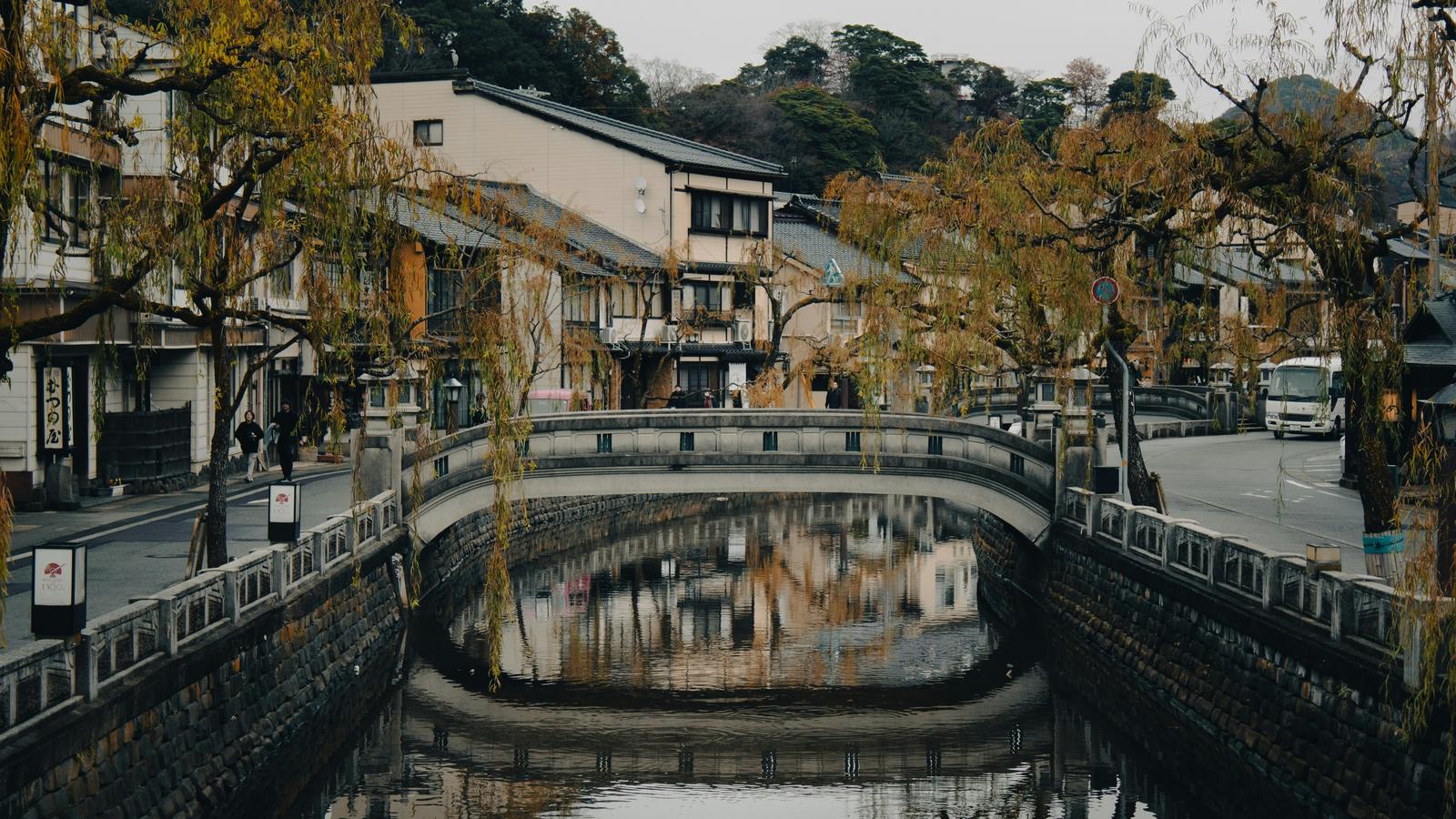 A stone bridge over the willow-lined canal at Kinosaki Onsen, with the town's inns reflected in the water