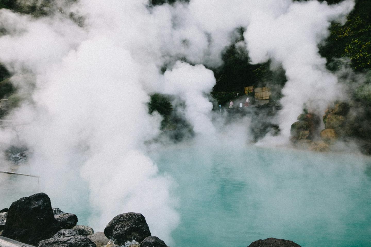 Thick steam rising from a turquoise hot spring pool among dark rocks and trees in Beppu