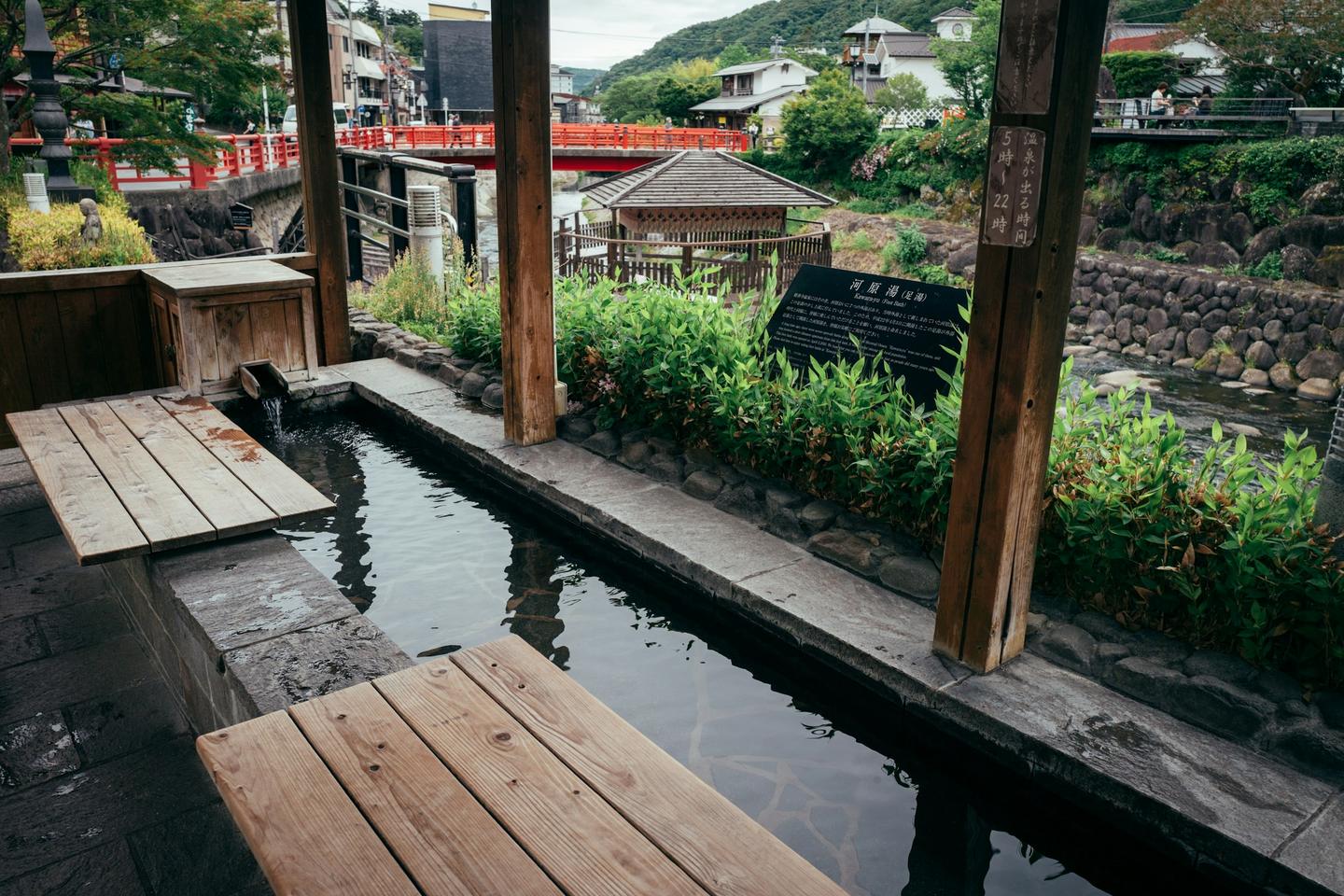 A covered outdoor foot bath with wooden benches beside a river in a hot spring town