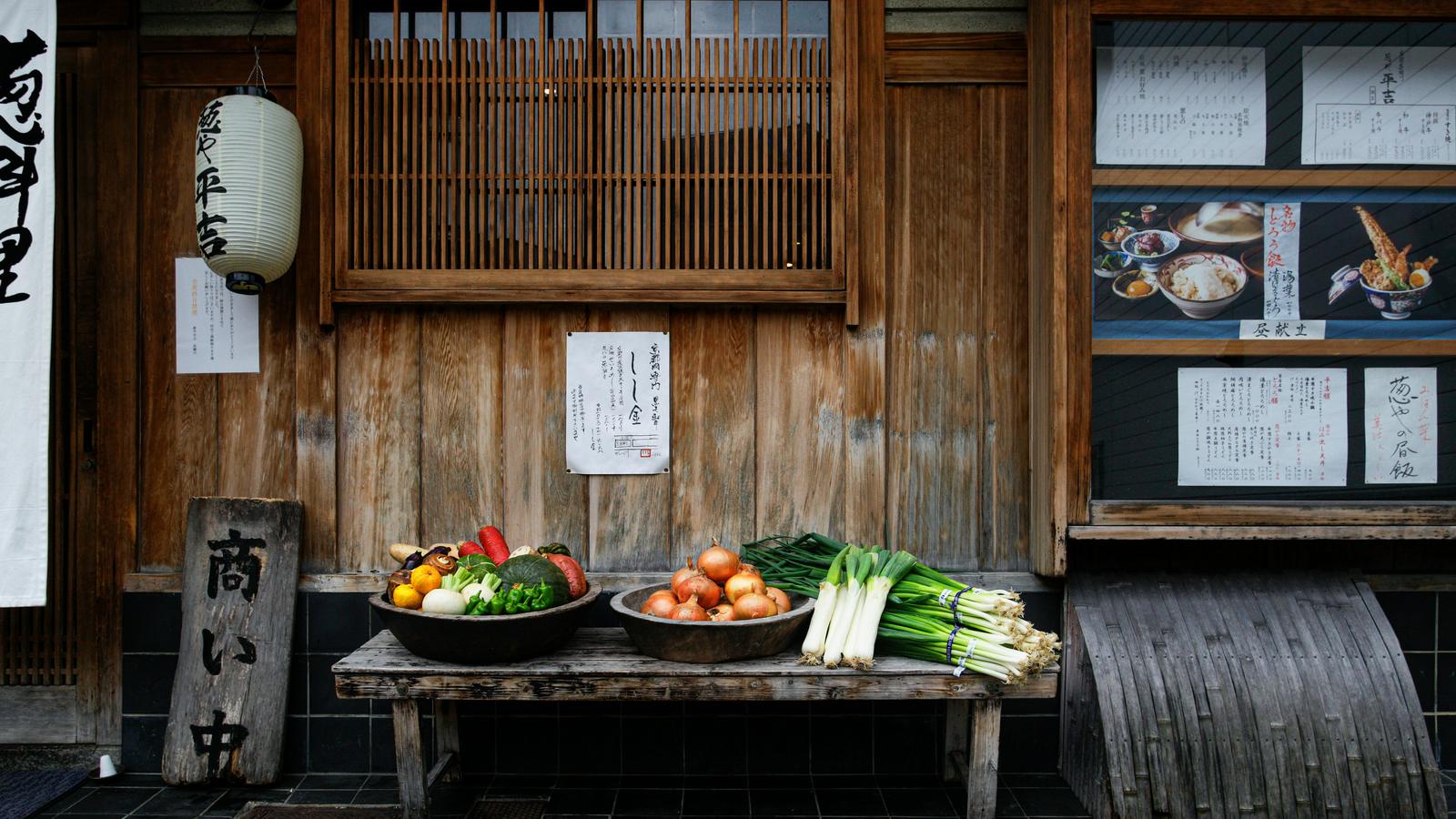 Baskets of fresh vegetables and leeks set out on a wooden bench in front of an old Japanese shop front