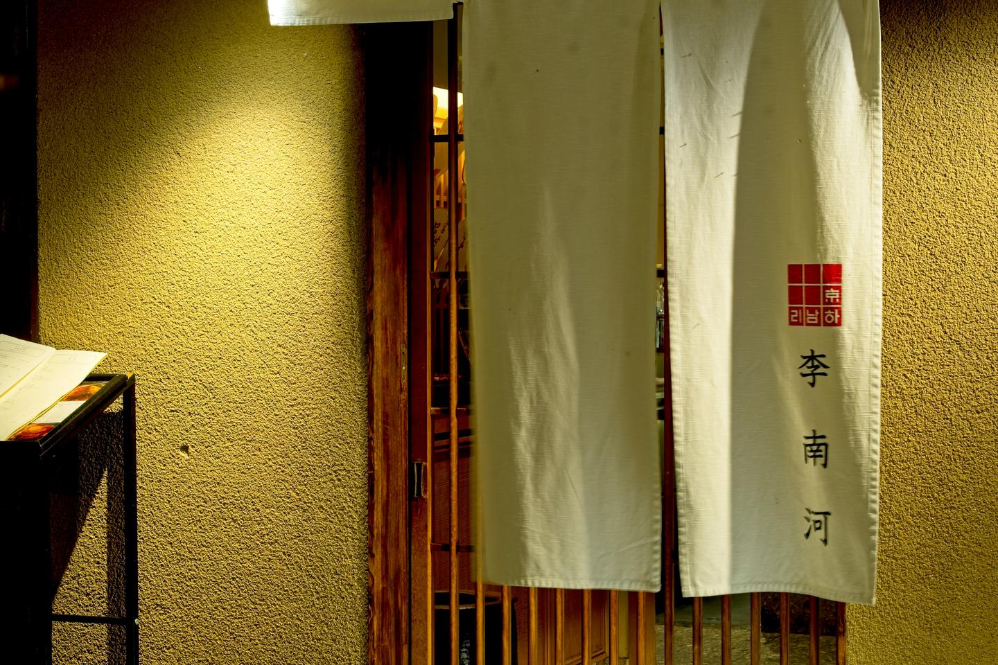 A pale noren curtain hangs over the lattice door of a restaurant at night, lit by a small lantern.