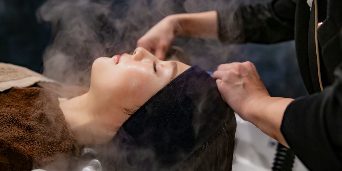 A guest lying back at a head spa basin while steam rises over her hair and a therapist works at her temples