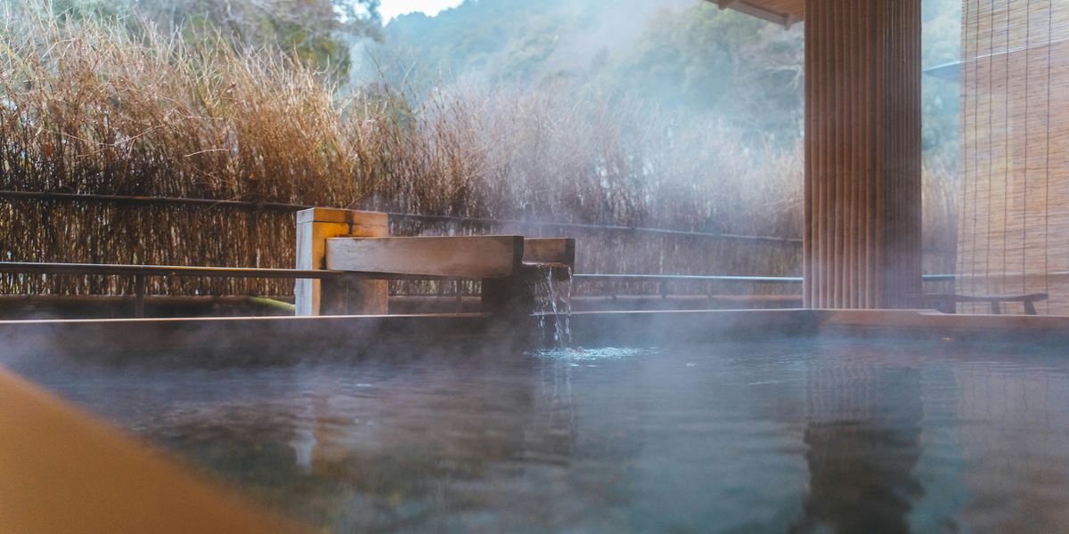 Steam rising from a ryokan's open-air bath in Kinosaki, with a wooden water spout, a brushwood fence and wooded hills beyond
