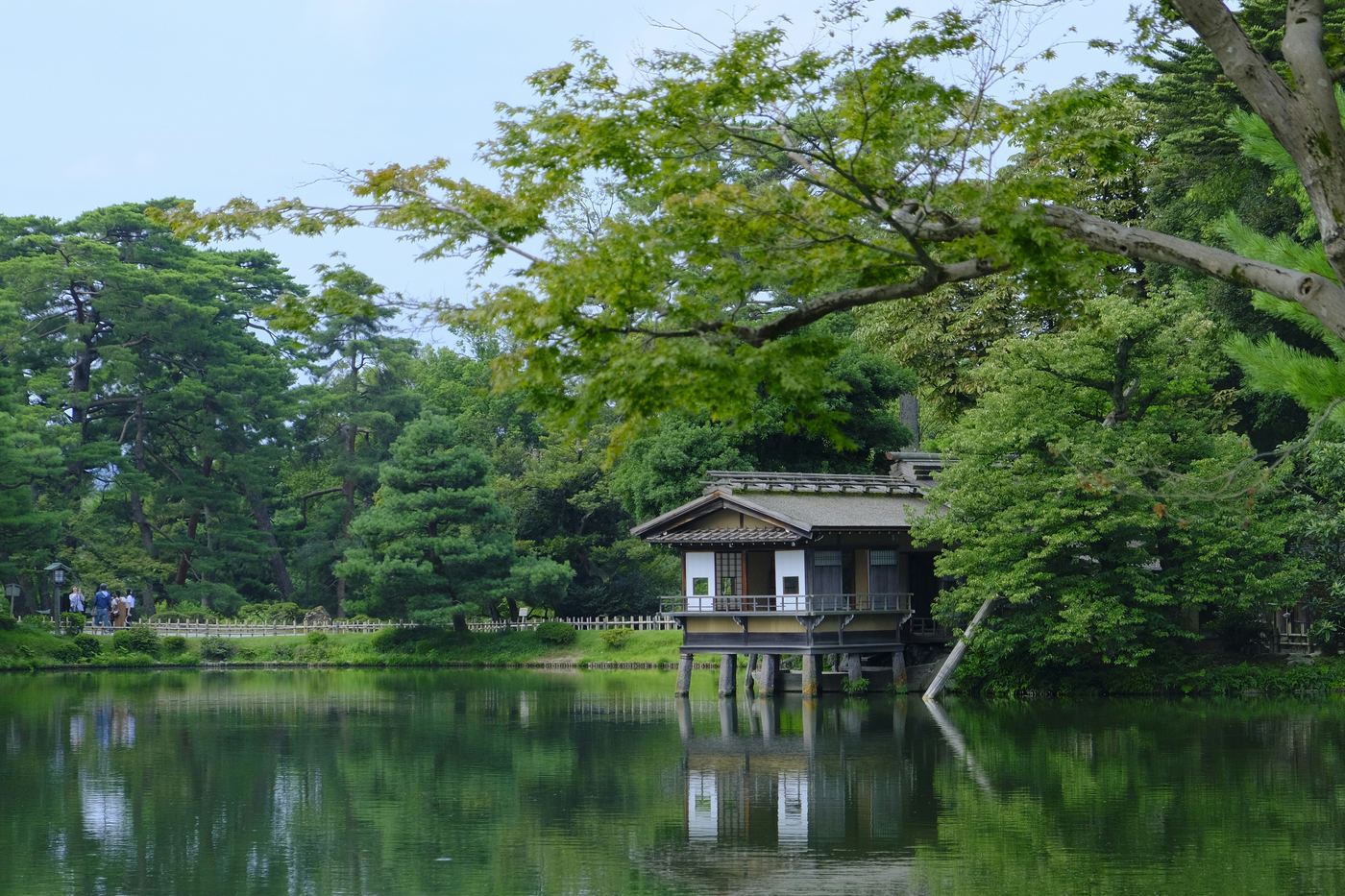 A wooden teahouse on stilts over a still pond in Kenroku-en, surrounded by tall green trees