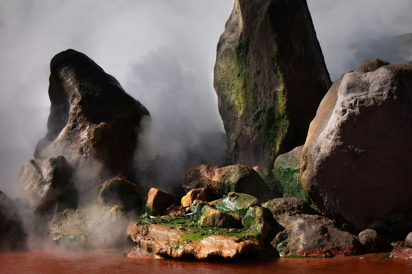Steam rising from a red hot spring pool among rocks in Beppu