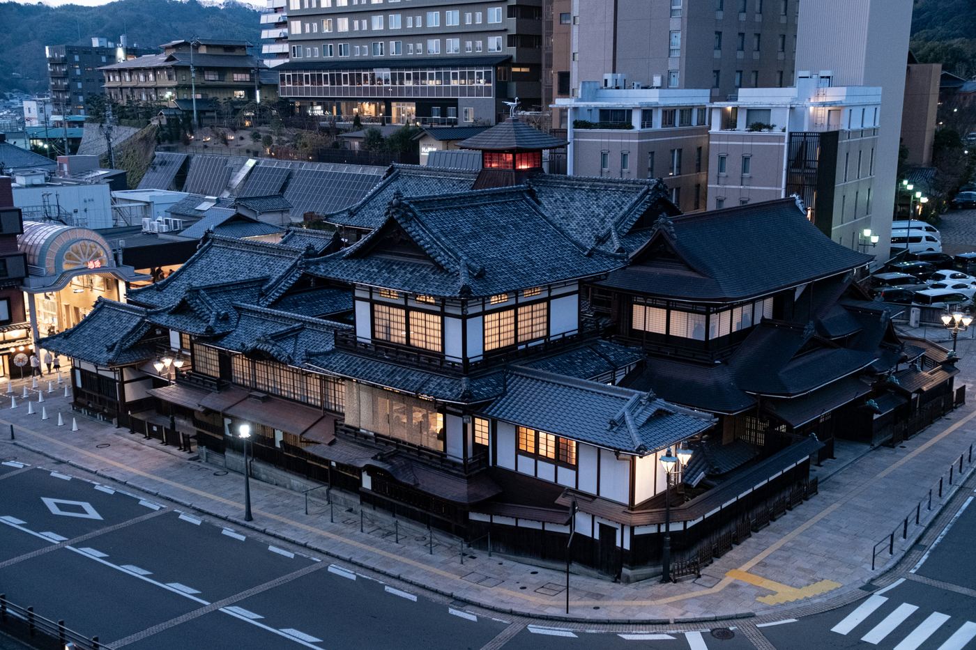 The wooden main bathhouse of Dogo Onsen at dusk, its tiled roofs layered above the lit street